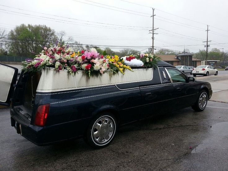 A polished black funeral hearse from the SRB Limo fleet adorned with beautiful, colorful floral arrangements on top, parked on a quiet street during an overcast day