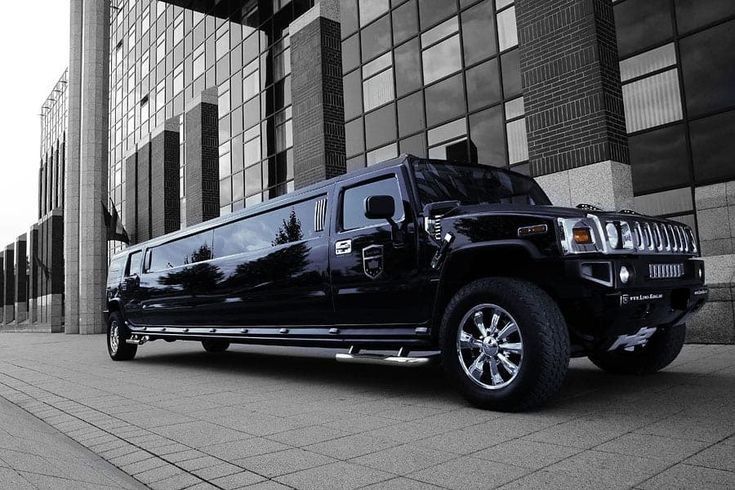 A side-angle black and white photograph of a long, polished black Hummer stretch limousine from the SRB Limo fleet parked on a sidewalk in front of a modern glass-windowed corporate building.