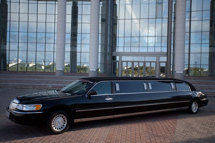 A side-profile view of a long, polished black stretch limousine from the SRB Limo fleet parked in front of a grand modern building with a massive glass facade and tall white pillars