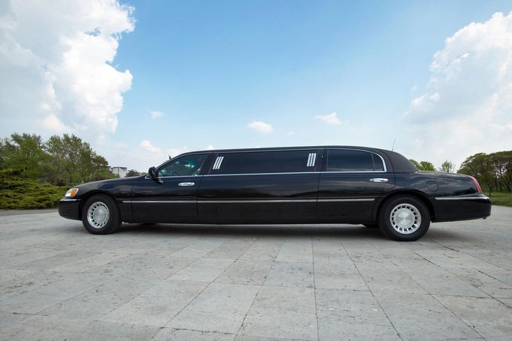 A side-profile view of a long, polished black stretch limousine from the SRB Limo fleet parked on a light stone pavement under a bright blue sky with white clouds