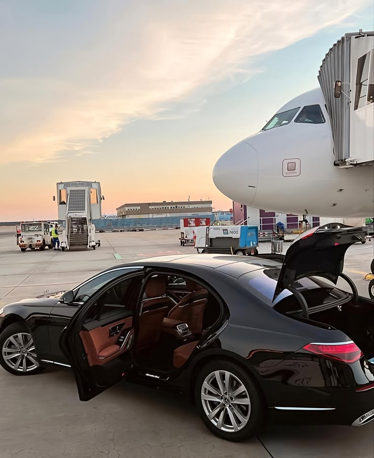 A high-end black Mercedes-Benz sedan from the SRB Limo fleet with an open door and trunk, parked directly on an airport tarmac next to a large commercial airplane during a golden sunset.