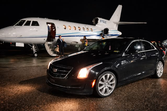 A polished black executive sedan from the SRB Limo fleet parked on a wet airport tarmac at night, positioned in front of a white private jet with its cabin lights on and stairs deployed.