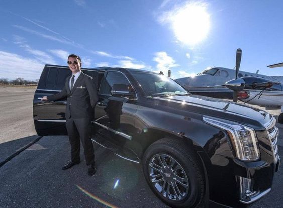 A professional chauffeur in a black suit and sunglasses opening the rear door of a polished black luxury SUV on an airport tarmac, with a private turboprop plane and a bright sun in the background.