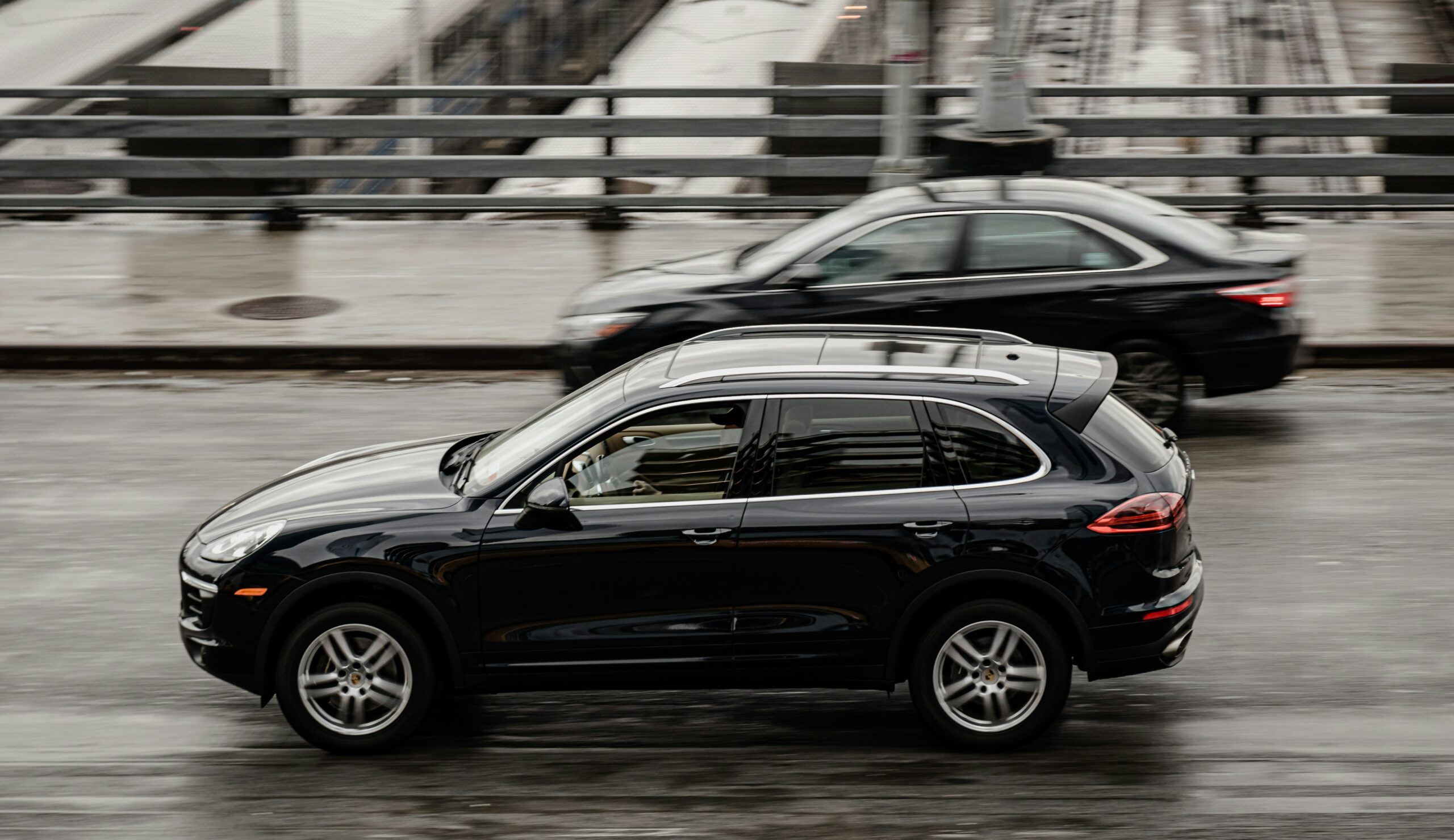A side-profile motion shot of a polished black luxury SUV from the SRB Limo fleet driving along a city road with a blurred background of urban structures