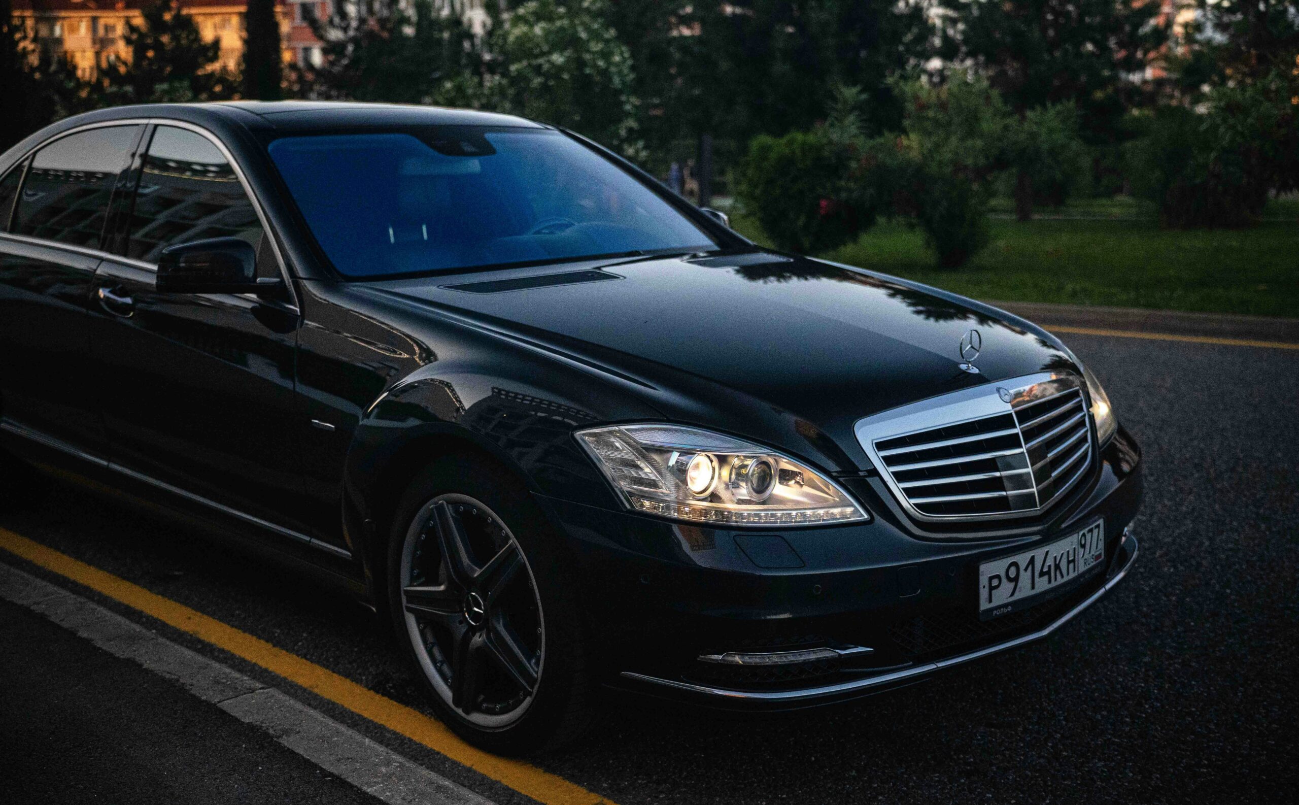 A close-up front view of a polished black Mercedes-Benz S-Class luxury sedan from the SRB Limo fleet parked on a street during twilight with its headlights illuminated.