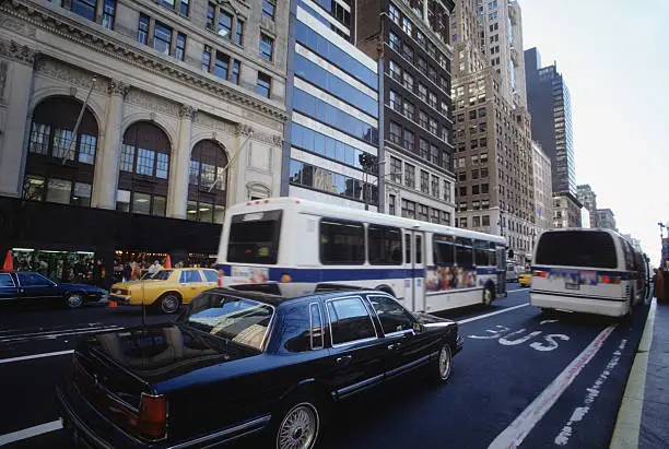 A rear-angle view of a polished black luxury sedan from the SRB Limo fleet driving through a busy city street lined with tall metropolitan buildings, alongside public buses and other city traffic
