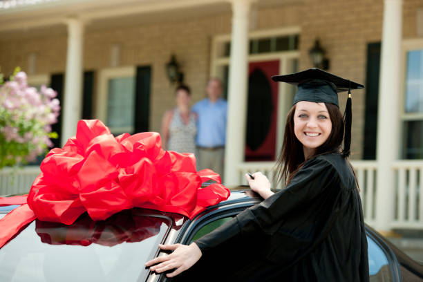A smiling young woman in a black graduation cap and gown standing next to a black car decorated with a large red gift bow, with her proud parents standing in the background on their house porch.