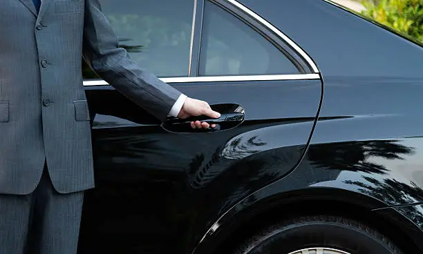 A professional chauffeur in a formal grey suit opening the rear door of a polished black luxury sedan for a passenger.