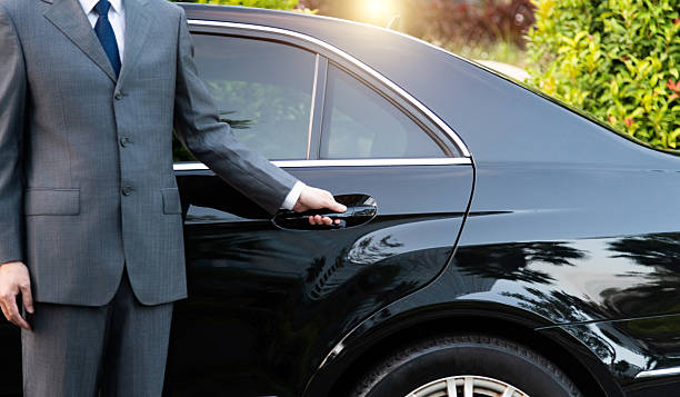 A close-up of a professional chauffeur in a formal grey suit opening the rear door of a polished black luxury sedan for a passenger.