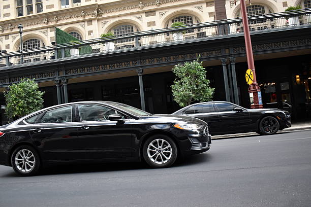 A side profile of a polished black executive sedan from the SRB Limo fleet driving on a clean city street, with another black luxury vehicle behind it and a classic building with arched windows in the background.