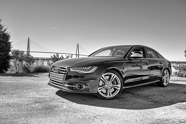 A front-angle black and white photograph of a sleek, polished executive sedan from the SRB Limo fleet parked on a gravel path, with a suspension bridge visible in the background under a clear sky.