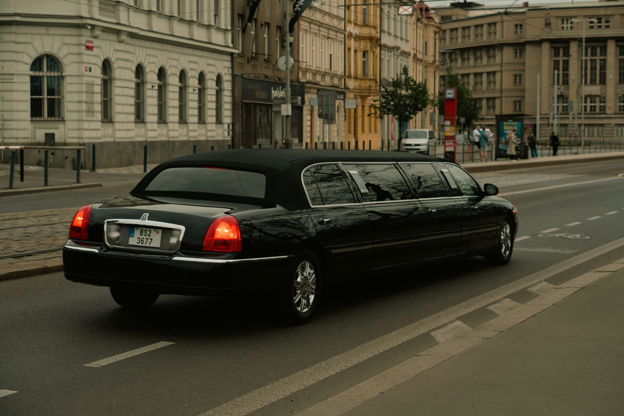 A rear-three-quarter view of a long, polished black stretch limousine from the SRB Limo fleet driving on a city street with historical buildings and a designated bike lane.