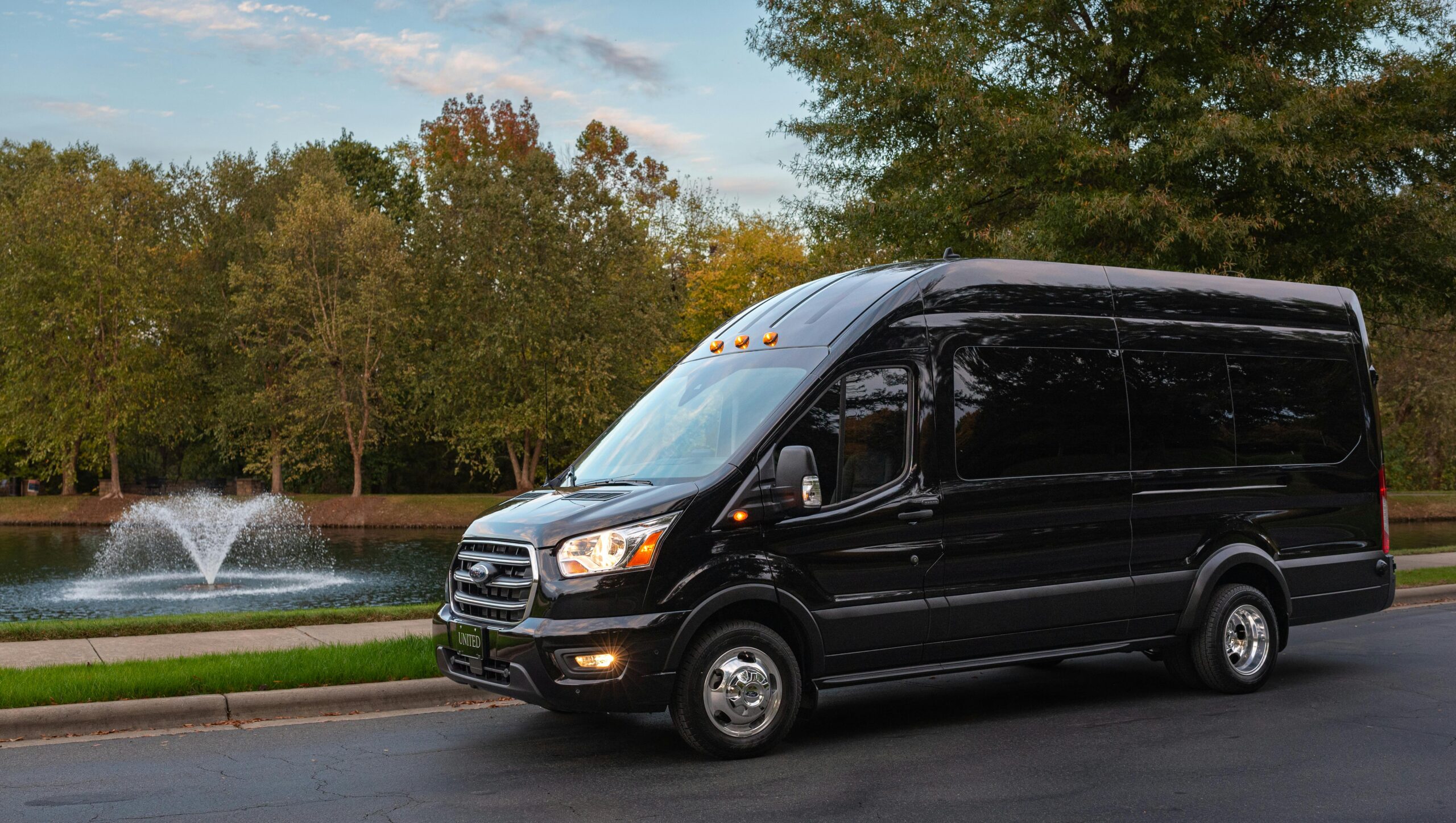A front-angle view of a sleek, high-roof black Ford Transit passenger van from the SRB Limo fleet parked on a quiet road next to a park with a fountain and green trees