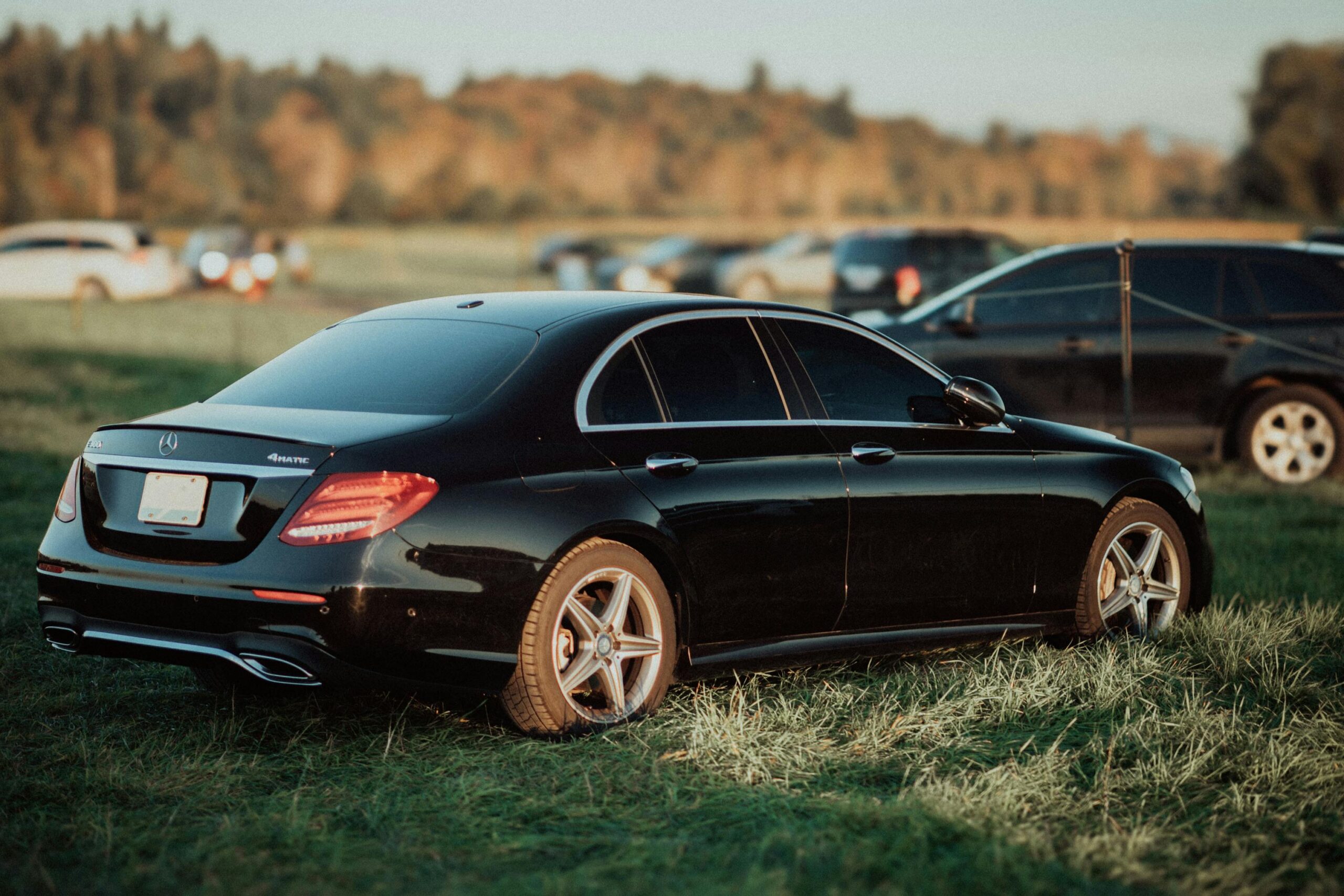 A side-rear view of a polished black Mercedes-Benz sedan from the SRB Limo fleet parked on a grassy field during an outdoor event at sunset.