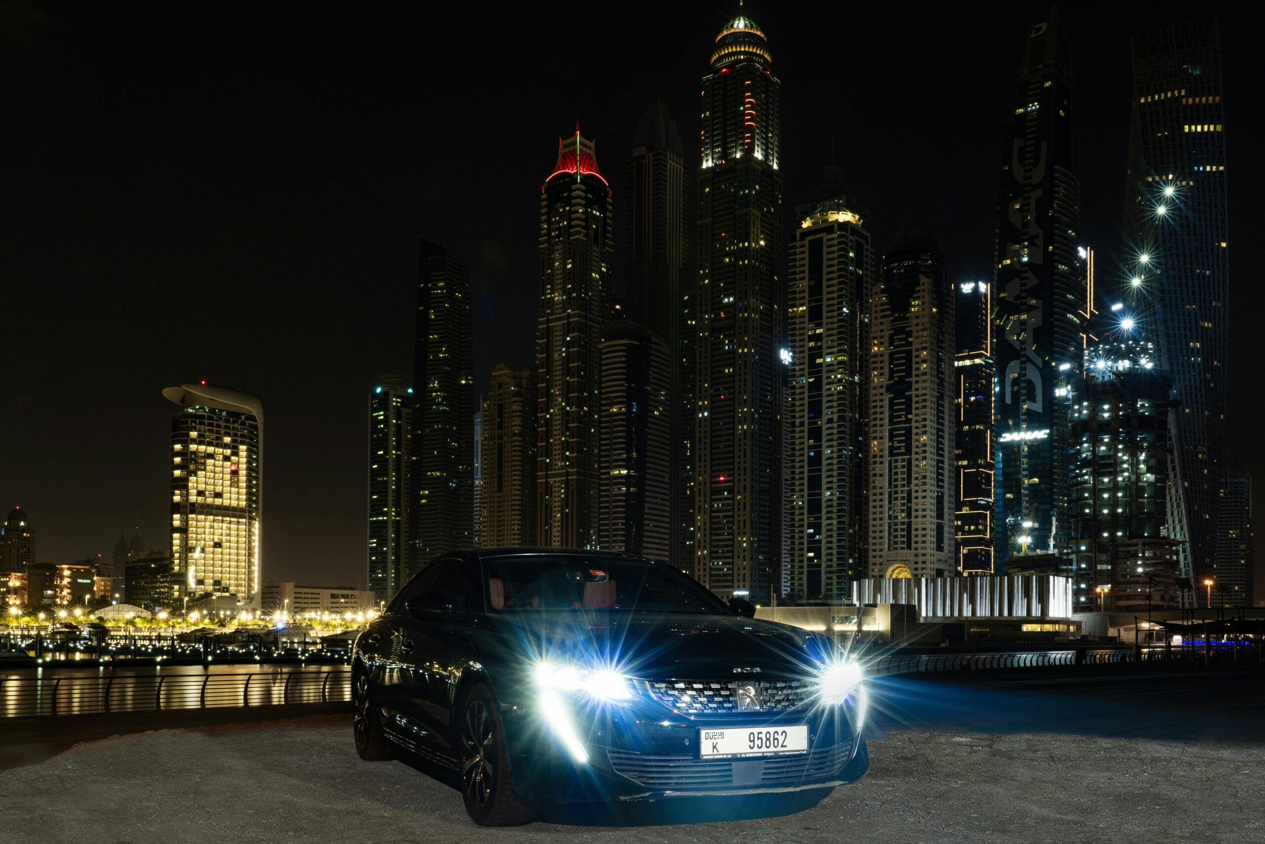 A front-angle view of a polished black luxury sedan from the SRB Limo fleet with bright headlights on, parked against a stunning backdrop of a lit-up city skyline and skyscrapers at night