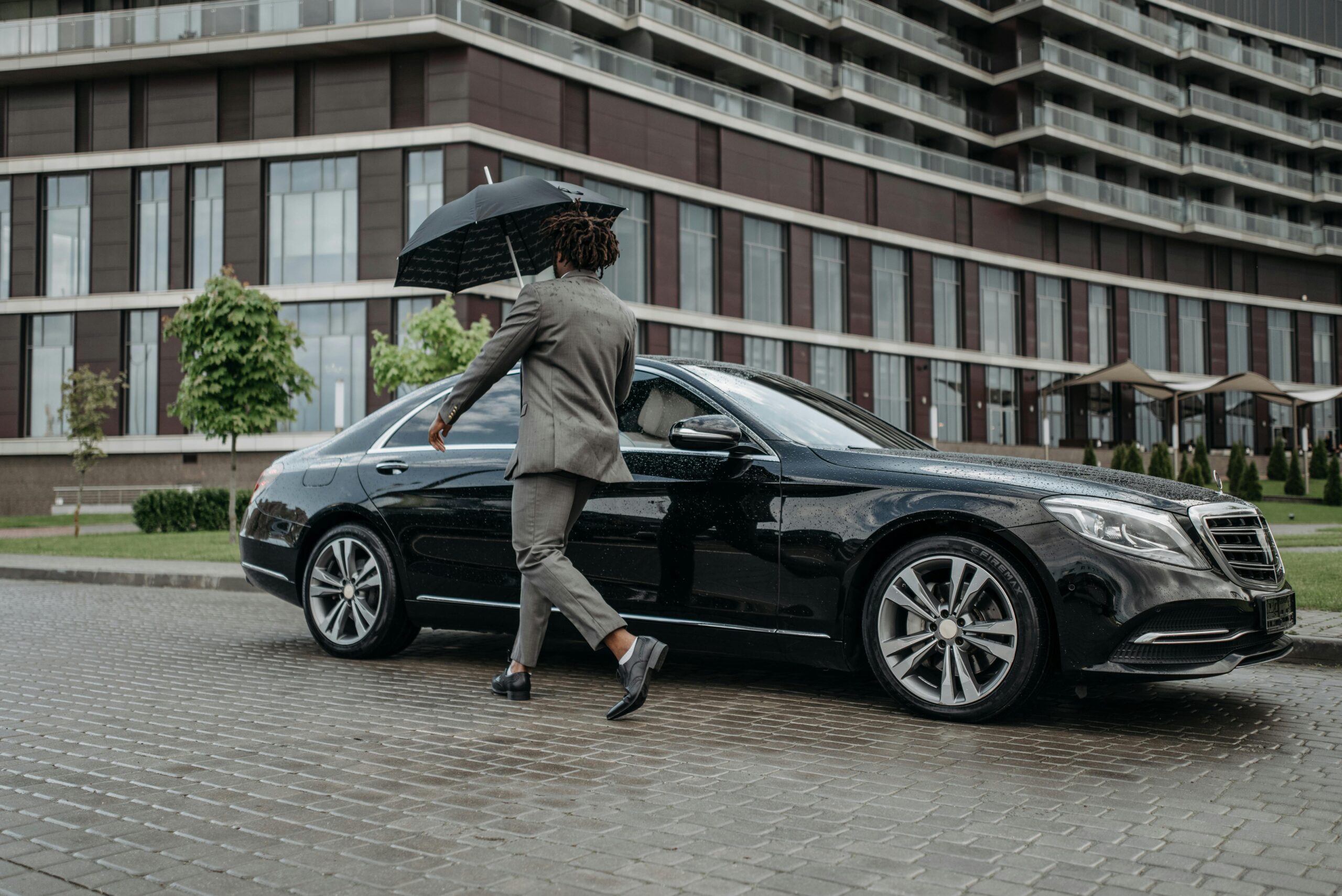 A professional in a suit with an umbrella approaching a luxury black sedan, representing our premium private group transportation in Sun City, AZ.