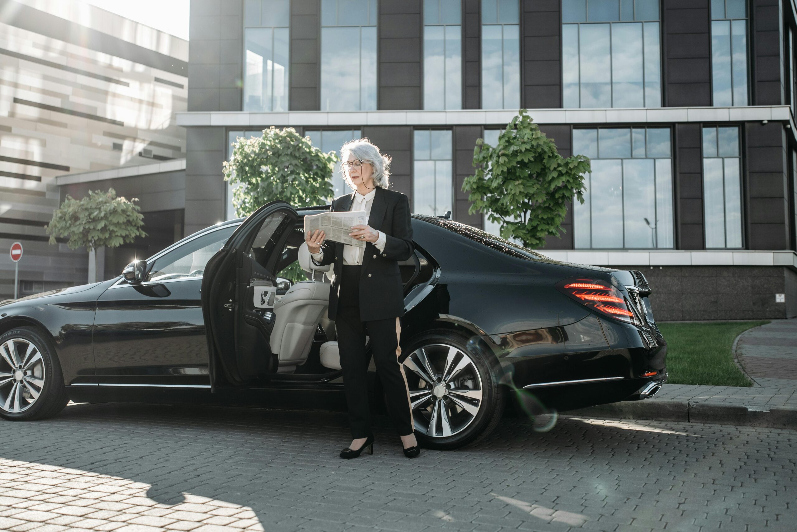 A professional businesswoman standing next to a black luxury Mercedes-Benz sedan from the SRB Limo fleet while reading a newspaper in front of a modern corporate office building.