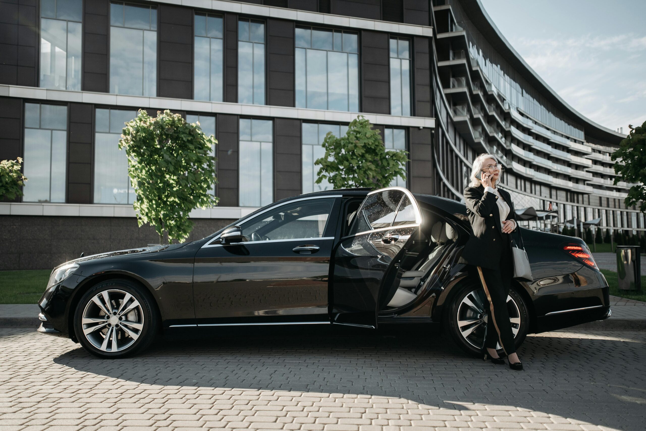 A side profile of a polished black luxury sedan with an open rear door, where a professional woman in a business suit is standing and talking on a phone in front of a modern corporate building.
