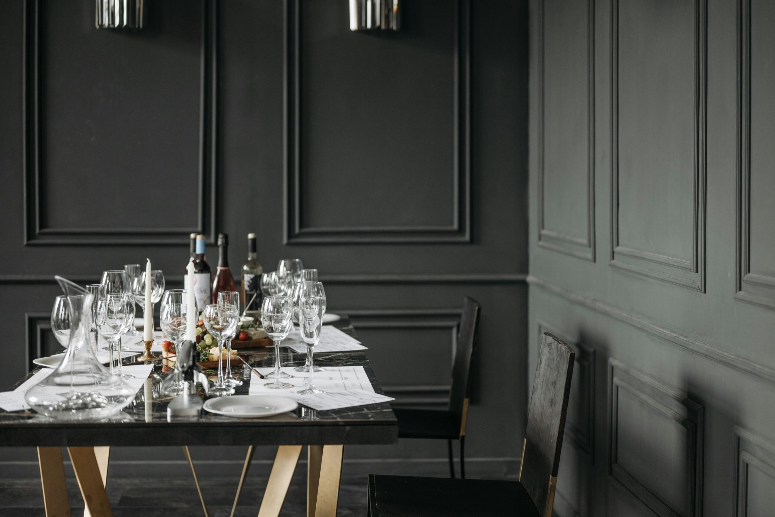 A sophisticated indoor dining setting featuring a dark marble table set with crystal glassware, wine bottles, and candles against elegant dark gray paneled walls.