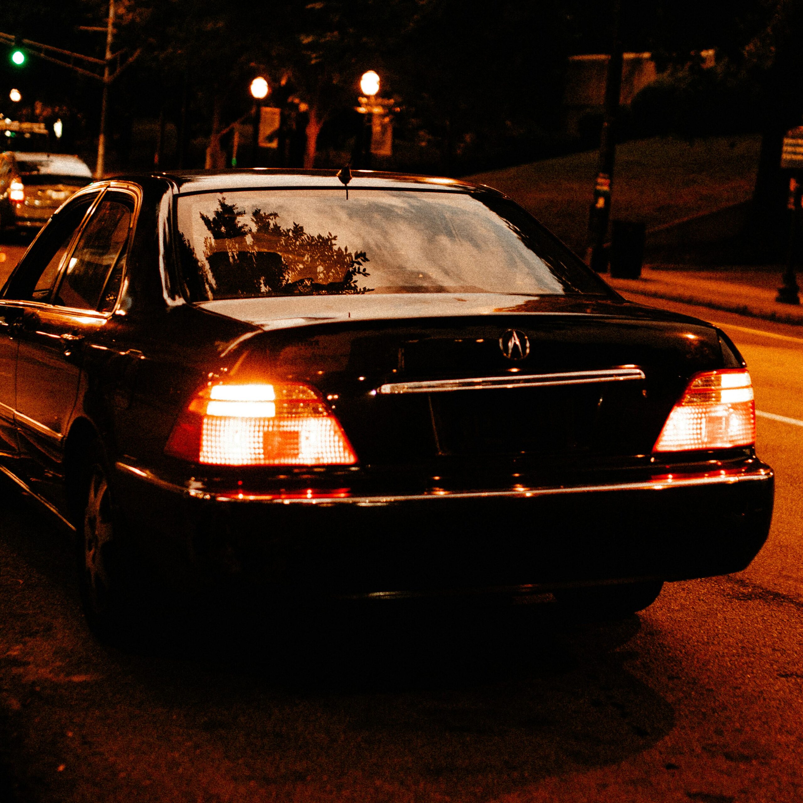 A close-up rear-angle view of a polished black luxury sedan from the SRB Limo fleet with its taillights illuminated, parked on a city street at night under warm street lighting