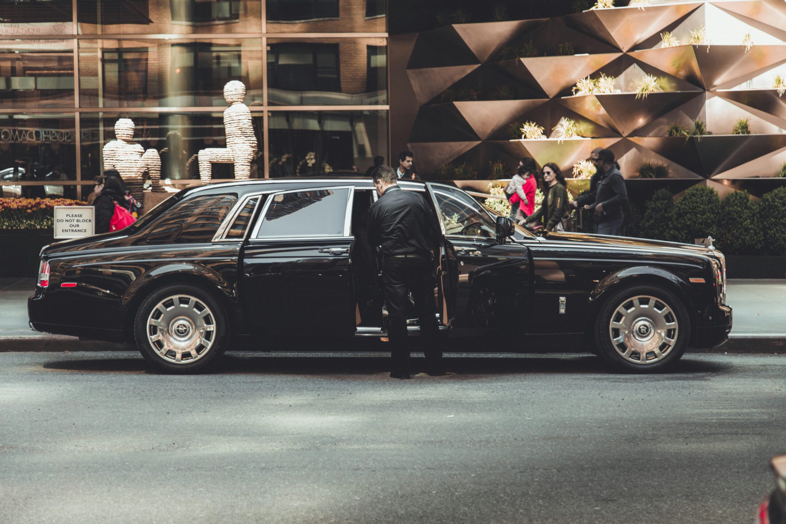 A side-profile view of a polished black ultra-luxury sedan parked on a city street, with a professional chauffeur in a black uniform opening the rear door for a passenger in front of a modern building with unique architectural decor.